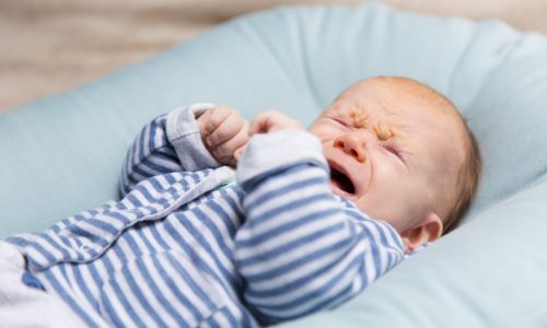 Crying red haired baby in blue and grey clothes, lying on soft pad. Portrait of cute little child in home interior. Discomfort concept