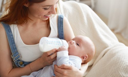 Sweet moment of happy young Caucasian mother smiling joyfully holding bottle of modified milk and feeding her adorable baby. People, lifestyle, childcare, maternity, love, family and happiness concept